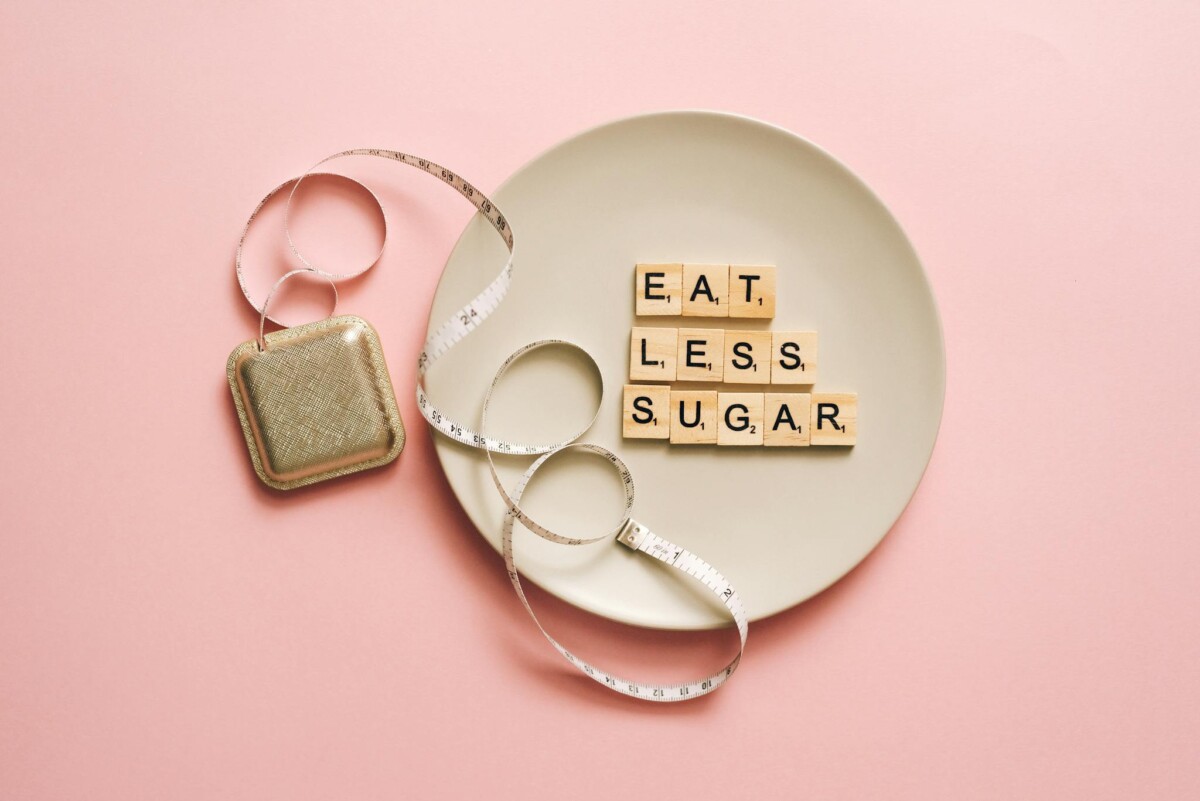 Flat lay of Scrabble tiles saying 'Eat Less Sugar' on a plate with a tape measure on a pink background.